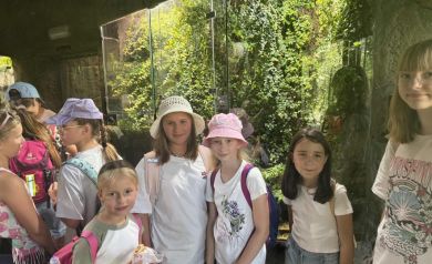 Na zdjęciu widać dziewczynki. Na pierwszym planie kilka z nich ustawia się do zdjęcia na tle przeszklonego pomieszczenia z roślinami.