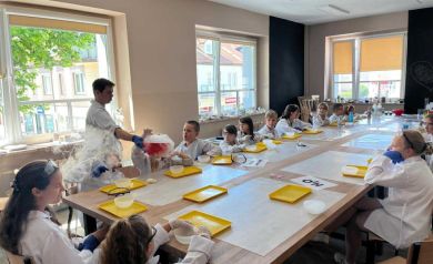 Dzieci wraz z prowadzącym podczas warsztatów chemicznych. Dzieci siedzą przy dużym stole, na którym można dostrzec głównie żółte tacki i okrągłe pojemniki. Mężczyzna prezentuje doświadczenie chemiczne, z którego wydobywa się bialo-czerwony dym. 