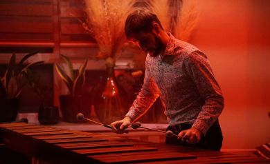 Muzyk grający na marimbie. W tle dominuje światło sceniczne w palecie odcieni czerownego koloru.