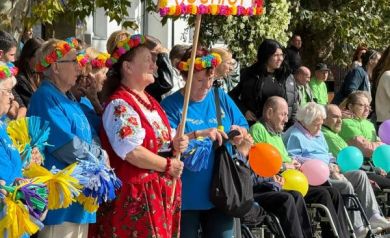 Uczestnicy wydarzenia, niektórzy poruszający się na wózkach inwalidzkich. Trzymają w dłoniach kolorowe balony i tabliczkę z napisem ,,Senior+'. Ubrani są w kolorowe stroje, na głowach mają kolorowe wianki.