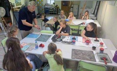 Na fotografii widać grupkę dzieci wraz z instruktorem. Dzieci siedzą na zielonych krzesłach przy stołach, na których znajdują się różne prace i przyrządy do ich wykonania. Obok dzieci stoi instruktor zajęć, który mówi do uczestników warsztatów. 