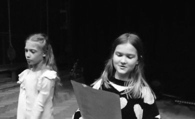 Na zdjęciu dwie dziewczynki, śpiewają. Jedna z dziewczynek trzyma w dłoniach kartkę z tekstem piosenki. Zdjęcie jest czarno-białe.