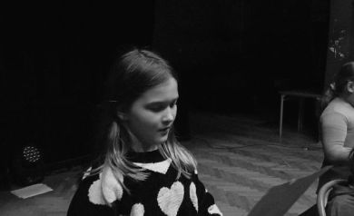 Na zdjęciu dziewczynka, która śpiewa. W dłoniach trzyma kartkę z tekstem. Zdjęcie jest czarno-białe. 