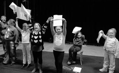 Na zdjęciu grupa dzieci unosząca kartki z tekstami piosenek do góry. Zdjęcie jest czarno-białe.