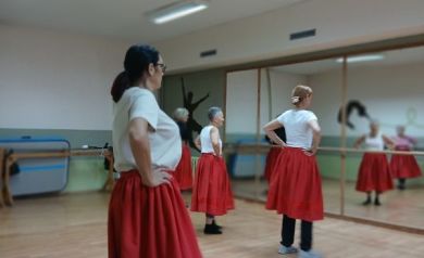 Na zdjęciu kilka seniorek ćwiczy rytmikę w sali tanecznej, stojąc przed lustrem w czerwonych spódnicach i białych bluzkach. Kobiety wykonują układ z rękami opartymi na biodrach.