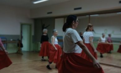 Na zdjęciu seniorki podczas zajęć rytmiki ćwiczą taneczny krok, trzymając czerwone spódnice i poruszając się po sali. W tle widać lustro oraz pozostałe uczestniczki wykonujące ten sam ruch.