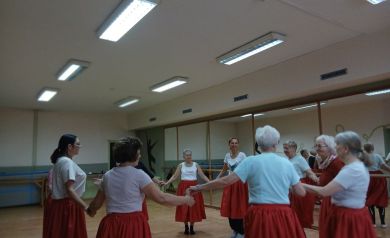 Na zdjęciu seniorzy biorą udział w zajęciach rytmiki, stojąc w kole w sali z lustrami i tańcząc w czerwonych spódnicach i jasnych bluzkach.