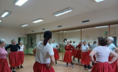 Na zdjęciu seniorzy biorą udział w zajęciach rytmiki, stojąc w kole w sali z lustrami i tańcząc w czerwonych spódnicach i jasnych bluzkach.