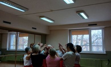Na zdjęciu grupa seniorek podczas zajęć rytmiki stoi w kręgu, unosząc ręce i tworząc wspólny taneczny gest. Wszyscy uczestnicy mają na sobie czerwone spódnice i ćwiczą w jasnej sali z lustrami.