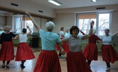 Na zdjęciu widać seniorki tańczące w parach, trzymające się pod pachę i poruszające się po sali w rytmie muzyki, wszystkie ubrane w czerwone spódnice. Atmosfera zajęć wydaje się pełna radości i swobody.