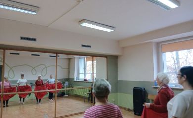 Na zdjęciu widać grupę seniorek ćwiczących rytmikę w sali tanecznej, trzymających czerwone spódnice podczas wspólnego układu. Zajęcia odbywają się przed dużą lustrzaną ścianą, dzięki której uczestniczki widzą swoje odbicia.