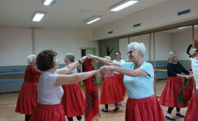 Na zdjęciu seniorki ćwiczą rytmikę w parach, tańcząc z kolorową chustą i uśmiechając się do siebie. Wszystkie uczestniczki mają na sobie czerwone spódnice i poruszają się po jasnej sali tanecznej.
