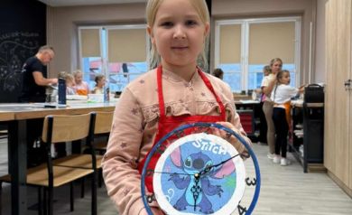Na zdjęciu dziewczynka w czerwonym fartuszku prezentuje wykonany przez siebie zegar. W tle widać innych uczestników warsztatów i sale, gdzie obywały się zajęcia. 