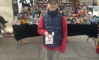 Chłopiec w czerwonej bluzie i granatowej kamizelce trzyma znalezioną torebkę niespodziankę w kształcie zajączka, w tle stoisko z szydełkowanymi maskotkami.