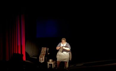 Na zdjęciu aktorka występująca na scenie. Siedzi na krześle i szyje. 