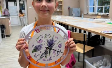 Dziewczynka ubtana w pomarańczowy fartuszek pozuje do zdjęcia z wykonanym przez siebie zegarem. W tle widać jednego z uczestników warsztatów i sale, gdzie obywały się zajęcia. 