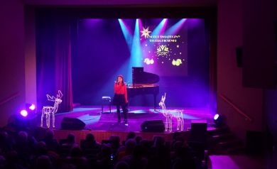 Publiczność ogląda młodą wokalistkę, która śpiewa na scenie w świetle fioletowych reflektorów.
Występ ma świąteczny klimat, poprzez wystrój na scenie.