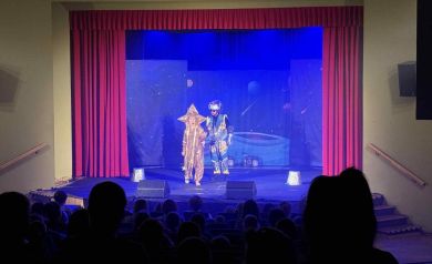 Na pierwszym planie publika. W oddali na scenie widać dwóch aktorów w przebraniach. Tło jest niebieskie, widać na nim kosmos. Dużą i dobrze widoczną dekoracją jest również czerwona kurtyna. 