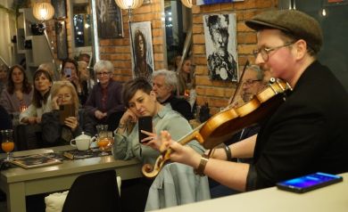 Publiczność nagrywa i ogląda występ muzyka. Widownia siedzi w restauracji pijąc napoje.