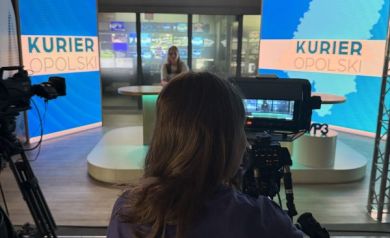 Na zdjęciu młoda dziewczyna w koszulce projektowej Równać Szanse 2025. Stoi za kamerą. W oddali studio nagrań Kuriera Opolskiego.  - powiększ