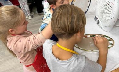 Dzieci suszące swoje zegary przy sprzęcie do tego przeznaczonym. Widać tam również inne przyrządy do tworzenia rękodzieła. 