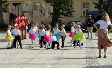 Na zdjęciu grupa przedszkolaków podczas swojego występu. Razem z nimi układ taneczny wykonuje kobieta, odwrócona przodem do dzieci. Przedszkolaki trzymają w dłoniach duże czerwone serca. Dziewczynki ubrane są w różnokolorowe spódniczki. Za nimi stoją w szeregu mażoretki, ubrane w czerwone sukienki. W tle widać również mężczyznę ubranego w okulary przeciwsłoneczne.