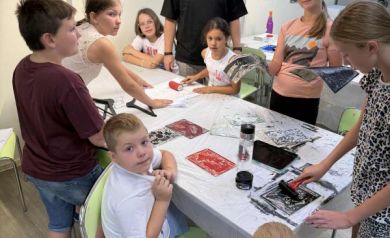 Uczestnicy zajęć wraz z instruktorem. Na stole, przy którym się gromadzą jest wiele obrazków. Na ścianie znajdują się różne obrazy i plakaty. 