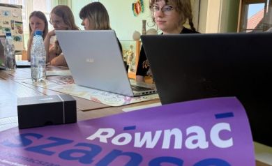Młodzież siedząca przy stole z laptopami podczas zajęć związanych z projektem. Widać również plakat promujący projekt.  - powiększ