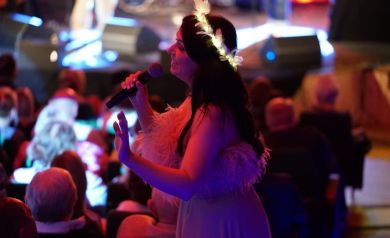 Na zdjęciu kobieta, która prawdopodobnie śpiewa do mikrofonu. Na głowie ma wianek z podświetlonych piór. W tle widać publiczność. 