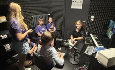 Na zdjęciu uczestnicy projektu w studio nagrań. Wokół nich wiele sprzętu muzycznego i do nagrań.  - powiększ