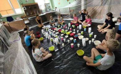 Grupa dzieci wraz z prowadzącą podczas zajęć z sensoplastyki. Na fotografii widać dużo różnych kubeczków z produktami spożywczymi potrzebnymi do zajęć. 