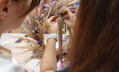 Na zdjęciu widać dwie kobiety uczestniczące w warsztatach florystycznych. Skupiają się na tworzeniu wianka z suszonych traw i drobnych kwiatów w odcieniach fioletu i bieli. Jedna z nich trzyma wstążkę, którą wplata w kompozycję, a druga uważnie przygląda się pracy. 