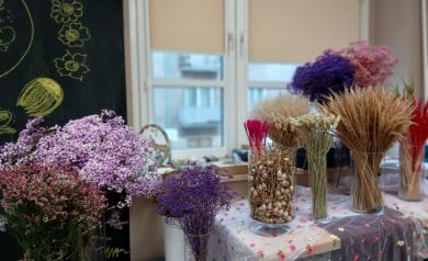 Na zdjęciu widzimy stół przygotowany do warsztatów florystycznych. Na nim ustawiono szklane wazony wypełnione różnorodnymi suszonymi roślinami – m.in. kłosami zbóż, trawami, kolorowymi kwiatami w odcieniach fioletu, różu, czerwieni i bieli. Stół przykryty jest materiałem ozdobionym drobnymi, kolorowymi kwiatkami, co nadaje całości lekkiego, wiosennego charakteru. W tle znajduje się czarna tablica z narysowanymi kredą kwiatami, a za stołem widać okna, przez które wpada naturalne światło. 