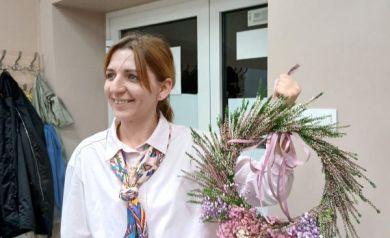 Na zdjęciu znajduje się uśmiechnięta kobieta, która prezentuje własnoręcznie wykonany wianek z kwiatów. Ubrana jest w jasnoróżową koszulę i kolorową, wzorzystą apaszkę. W dłoni trzyma okrągły wianek ozdobiony wrzosem, różowymi i fioletowymi kwiatami oraz różową wstążką. Kobieta stoi na tle drzwi z szybami, a w tle widać również wieszak z kilkoma kurtkami.