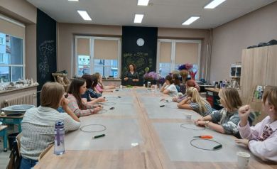 Na zdjęciu uczestniczki warsztatów siedzą przy stołach i słuchają prowadzącej, która tłumaczy kolejne etapy tworzenia wianków. Na stołach leżą obręcze i narzędzia, a w tle widać kolorowe suszone kwiaty przygotowane do pracy.