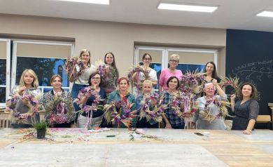 Na zdjęciu widać grupę kobiet pozujących wspólnie po zakończonych warsztatach. Każda z nich trzyma w rękach własnoręcznie wykonany wianek z suszonych kwiatów i traw, ozdobiony kolorowymi wstążkami. Uczestniczki stoją w dwóch rzędach, uśmiechając się do zdjęcia. Na stole przed nimi leżą resztki kwiatów i materiałów używanych podczas zajęć. 