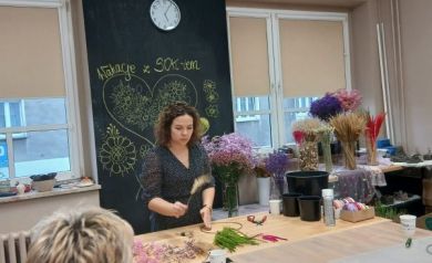 Na zdjęciu widać prowadzącą warsztaty florystyczne, która stoi przy stole i pokazuje uczestniczkom, jak przygotowywać rośliny do plecenia wianków. Przed nią leżą różnokolorowe kwiaty i trawy, a obok znajdują się sekatory oraz inne potrzebne materiały. Na pierwszym planie siedzą dwie uczestniczki, uważnie obserwując instrukcję. W tle widoczna jest tablica z rysunkami kwiatów i napisem „Wakacje z SOK-iem”, a także stoły z ustawionymi bukietami suszonych roślin i materiałami do dekoracji.