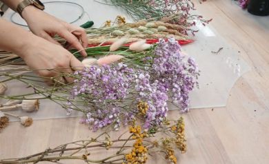 Na zdjęciu widać stół pełen suszonych kwiatów i roślin w różnych kolorach – fioletowych, różowych, żółtych i zielonych. Uczestnicy warsztatów, których dłonie są widoczne, przygotowują kompozycje, układając rośliny w małe wiązki, które później będą wykorzystane do tworzenia wianków. Na stole leżą także nożyczki, sekatory i metalowe obręcze do mocowania dekoracji.