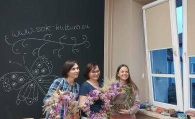 Na zdjęciu widzimy trzy uśmiechnięte kobiety stojące obok siebie i prezentujące własnoręcznie wykonane wianki z suszonych kwiatów i traw. Panie pozują na tle tablicy z rysunkiem motyla oraz napisem „www.sok-kultura.eu”. Po prawej stronie znajduje się okno z widokiem na zewnątrz, a na parapecie ustawione są różne ozdoby i drobiazgi.