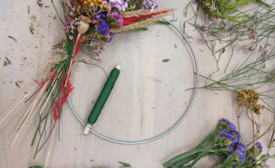 Na zdjęciu widać początek tworzenia wianka florystycznego. Na metalowej obręczy została już przymocowana pierwsza część kompozycji z suszonych i barwionych roślin – widoczne są fioletowe, żółte, czerwone oraz zielone gałązki i kwiaty. Obok leży zielony drut florystyczny, używany do mocowania roślin. Na stole porozkładane są różne suszone kwiaty i trawy w żywych kolorach: fioletowym, czerwonym, żółtym i zielonym. 