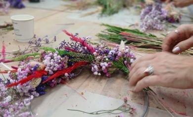 Na zdjęciu widzimy fragment warsztatów florystycznych. Na pierwszym planie znajduje się metalowa obręcz, do której uczestniczka przyczepia kolorowe kwiaty i trawy, tworząc wianek. Dominują barwy fioletowe, różowe, czerwone i zielone. W tle widać inne osoby zajęte podobną pracą – przy długim stole pełnym suszonych roślin, narzędzi i akcesoriów.