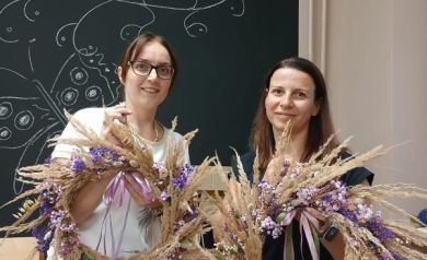 Na zdjęciu stoją dwie kobiety, każda trzyma w rękach własnoręcznie wykonany wianek z suszonych traw i kwiatów. Wianki są duże, ozdobione fioletowymi i różowymi kwiatami oraz wstążkami. Obie kobiety uśmiechają się do zdjęcia, a w tle widać czarną tablicę z rysunkiem motyla i adresem strony internetowej. Atmosfera zdjęcia jest radosna i pokazuje dumę z efektu pracy podczas warsztatów florystycznych.