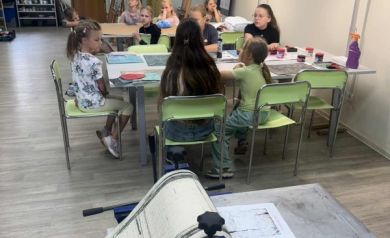 Przy stoliku na drugim planie dzieci siedzące na zielonych krzesłach, na pierwszym planie zaś maszyna używana podczas zajęć. 