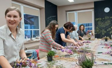 Na zdjęciu widać grupę kobiet pracujących przy długim stole, podczas warsztatów tworzenia wianków z suszonych kwiatów. Stół pokryty jest gałązkami, kwiatami i narzędziami florystycznymi, a uczestniczki skupiają się na swoich kompozycjach. Na pierwszym planie jedna z kobiet uśmiecha się, trzymając w dłoniach niemal gotowy wianek. W tle, na tablicy kredowej, widnieją rysunki i napis „Wakacje z SOK-iem”. 