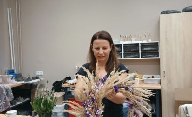 Na zdjęciu znajduje się kobieta trzymająca w dłoniach duży, efektowny wianek wykonany z suszonych traw i kwiatów w odcieniach fioletu. Stoi przy stole, na którym leżą gałązki, nożyczki florystyczne, sznurek i inne akcesoria do tworzenia wianków. W tle stoją doniczki z wrzosami oraz różne materiały florystyczne. 