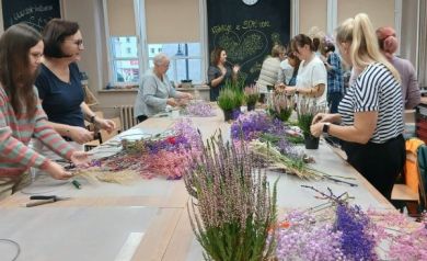Na zdjęciu widać uczestniczki warsztatów florystycznych podczas pracy nad wiankami. Kobiety stoją przy dużym stole, na którym rozłożone są kolorowe kwiaty, gałązki wrzosów i traw, a także narzędzia takie jak sekatory i druty do formowania wianków. Atmosfera jest twórcza i pełna skupienia – każda z osób zajmuje się swoim projektem. W tle widać prowadzącą oraz tablicę ozdobioną rysunkami i napisem, a cała sala jest jasna i przyjazna, z dużymi oknami wpuszczającymi światło dzienne.