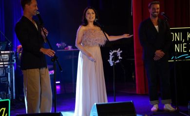 Na scenie artyści - kobieta ubrana w długą, jasną suknię i dwaj mężczyźni. Wszyscy się uśmiechają. Kobieta wskazuje prawą ręką na jednego z mężczyzn. 