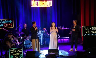 Na zdjęciu muzycy występujący na scenie. Widać również ledy z różnymi napisami. 