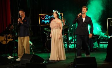 Dwaj mężczyźni i kobieta śpiewają do mikrofonów. Za nimi sprzęt muzyczny i dwaj muzycy. Tło stanowią zielone kolory i ciężki dym. 