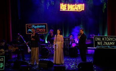 Wokaliści na scenie, śpiewają do mikrofonów. Widać również napisy led, sprzęt muzyczny, muzyków grających na instrumentach. Tło stanowią niebiesko-fioletowe światła oraz ciężki dym.